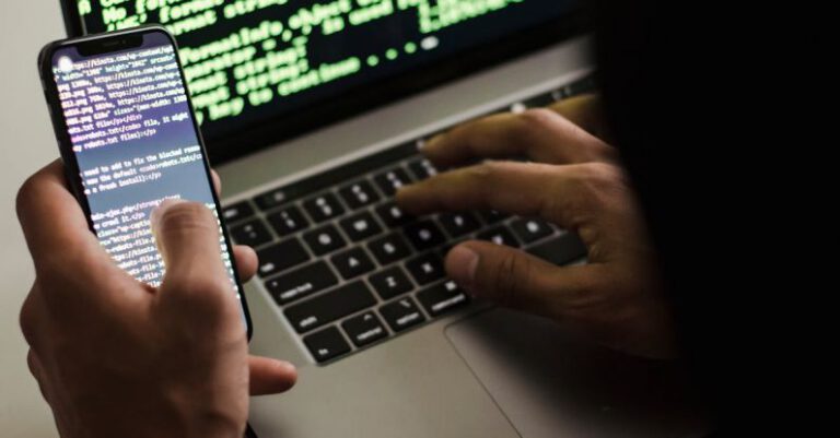 Code Optimization - Unrecognizable hacker with smartphone typing on laptop at desk